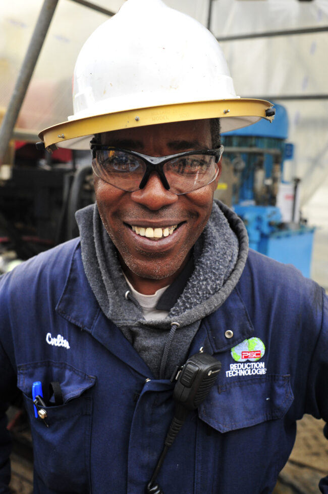Smiling oil rig worker wearing a white hard hat and safety glasses, with a blue work jacket, and a radio, demonstrating proper PPE. The worker's jacket features the Reduction Technologie logo. - David Joel Photographer