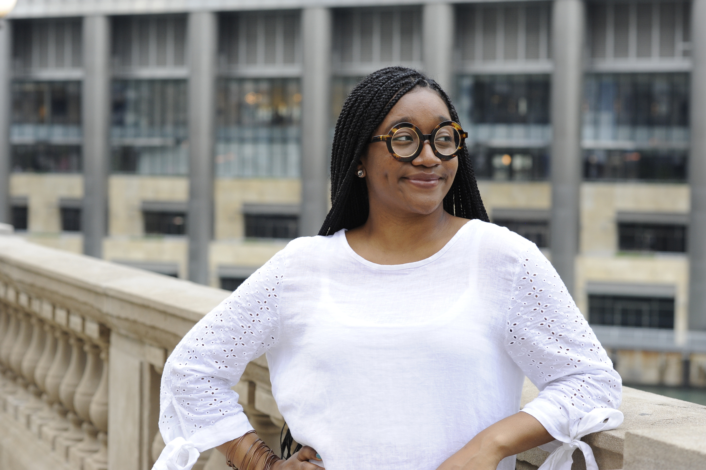 Portrait of a black woman in a white blouse and oversized circular glasses, standing on a balcony and smiling with a modern city building behind her. Portrait by David Joel Photography