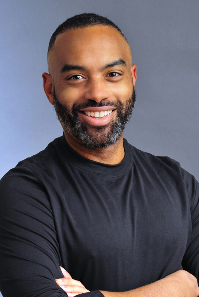 A smiling Black man with a neatly trimmed beard and short, dark hair. He wears a black long-sleeved shirt and has his arms crossed, radiating happiness and well-being. The background is a soft gradient of blue and grey. - David Joel Photographer